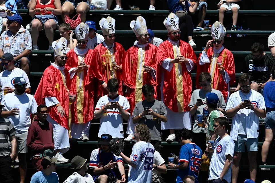 Six fans wearing red and gold robes and white mitres with White Sox logos in the stands among other baseball fans