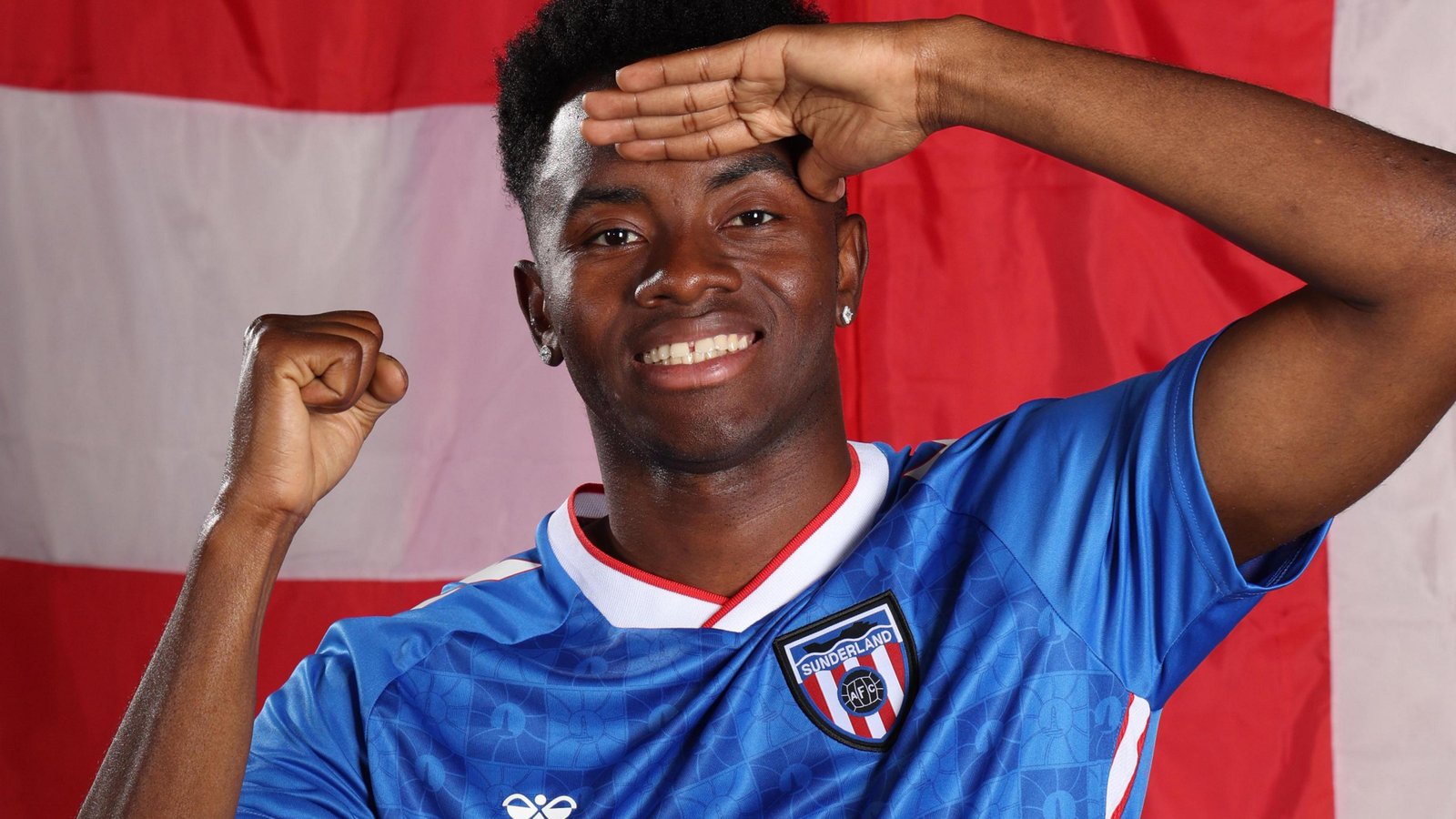 Simon Adingra, wearing a blue Sunderland away shirt, holds his right hand up in a fist and salutes with his left hand while standing in front of a red and white backdrop