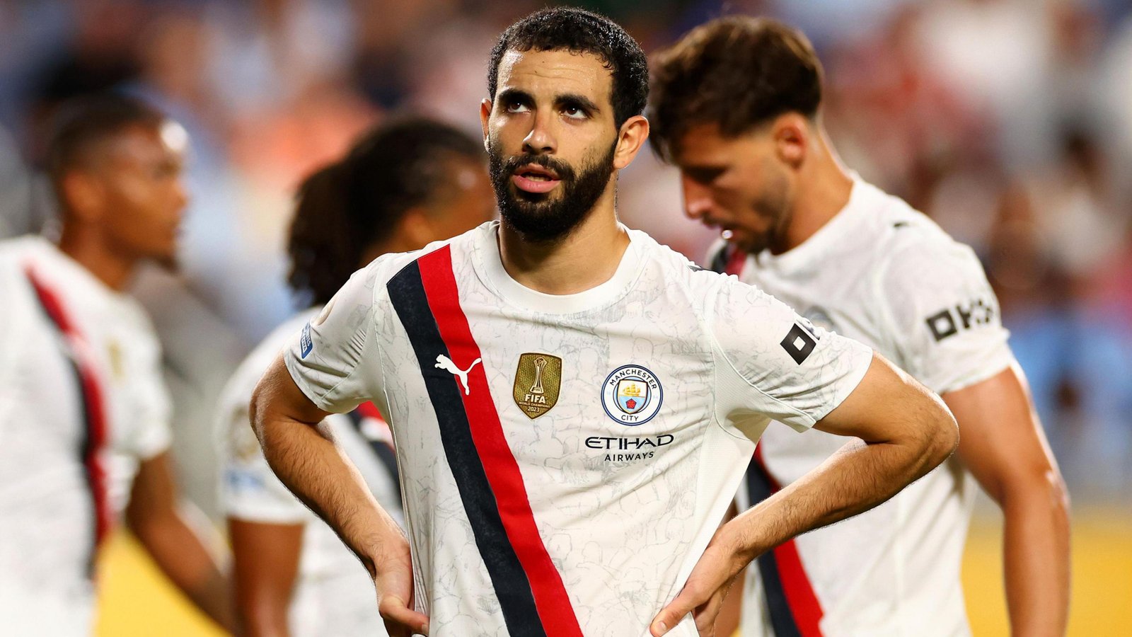 Rayan Ait-Nouri, wearing a cream Manchester City shirt with a black and red diagonal sash stripe, stands with his hands on his hips and raises his eyes up during a football match, with three team-mates visible but blurred behind him