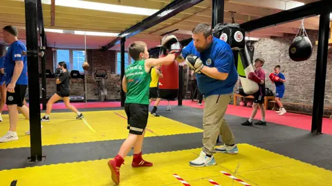 A boy in a green vest, black shorts  and red boots punches a hand pad being held by a bearded man in a blue t-shirt and cream trousers, with other young boxers in the background
