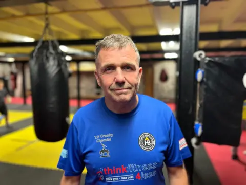 A grey-haired man, wearing a blue t-shirt, looks at the camera. Behind him is a boxing gymnasium