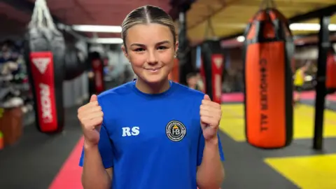 BBC A girl with blonde-streaked hair, wearing a blue t-shirt, smiles at the camera. Behind her are punchbags