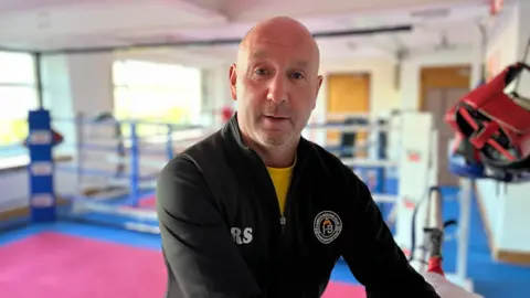 A bald-headed man, wearing a black top, look at the camera. Behind him is a boxing ring