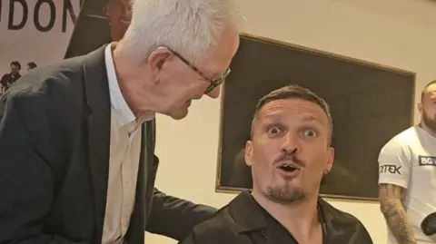Paul Gilfeather Frank Gilfeather speaking to the heavyweight champion Oleksandr Usyk, Frank is wearing a black suit with a white shirt laughing and Usyk is wearing a black shirt while looking at the camera surprised.