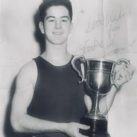 Paul Gilfeather A vintage black and white photo of a young Frank Gilfeather wearing a dark tank top holding a trophy