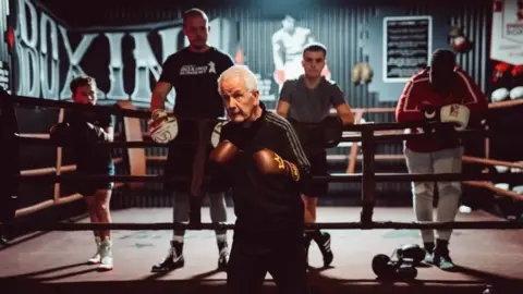 Paul Gilfeather Frank Gilfeather is just outside the ring in a boxing stance wearing a black tracksuit with a white trim. He is also wearing brown boxing gloves with golden crowns on them and behind him are four onlooking students who are standing inside the ring leaning on the ropes.