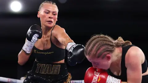 Getty Images Tiah-Mai in the ring, she black boxing gloves on, black short and top, her is braided. The other boxer is wearing red gloves and is crouching down in the right hand side.