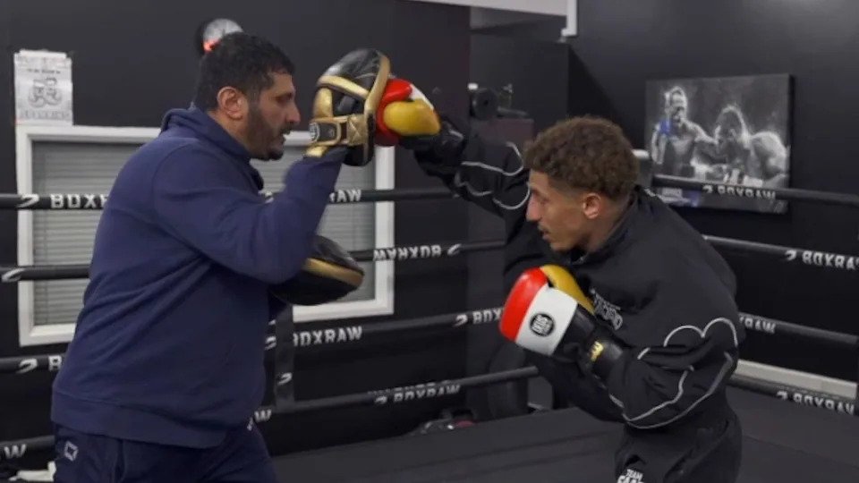 Saad is wearing a black tracksuit and red, white, yellow gloves. He is punching towards gold and black pads that his dad is holding. He is wearing a navy blue tracksuit. 