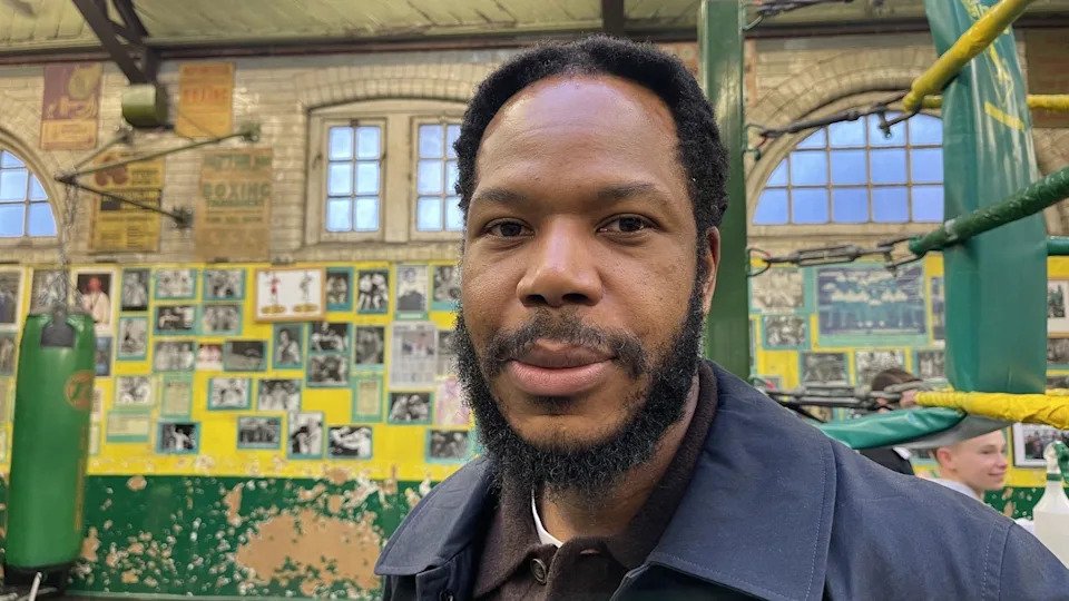 A man with dark hair and a beard stands in a boxing club. He is wearing a navy coat and a brown polo top with white T-shirt underneath. There are posters on the walls behind him and arched windows above.
