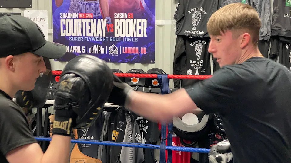 Two young men are in a boxing ring. One wears black cushioned hand pads, a black t-shirt and black cap. The other has short brown hair, a black t-shirt, black boxing gloves and is hitting the cushioned pads. In the background, there are many t-shirts and boxing posters pinned to the walls of the gym.