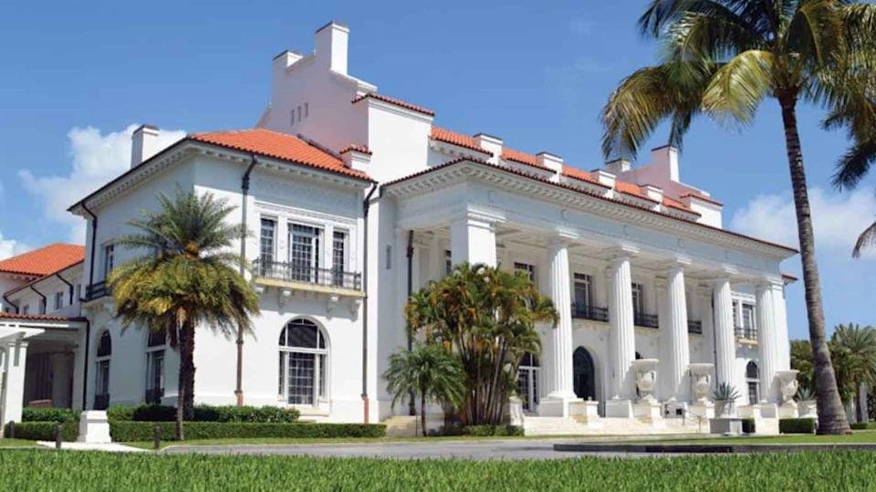 Elegant white mansion with red-tiled roof, grand columns, and large windows, surrounded by palm trees and manicured lawn under a bright blue Boca Raton sky—perfect for relaxing after a golf trip.