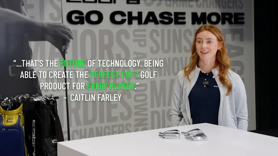 A woman with long red hair sits at a white table with two Cobra 3DP Irons golf club heads on it, smiling. Beside her is a rack of golf clubs. A quote about golf technology is displayed on the wall behind her.