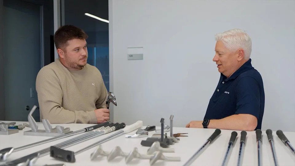 Two men talk at a white table displaying various golf club heads, shafts, and Cobra 3DP Irons in an office setting. One man holds a golf club while the other listens, both appearing engaged in conversation.