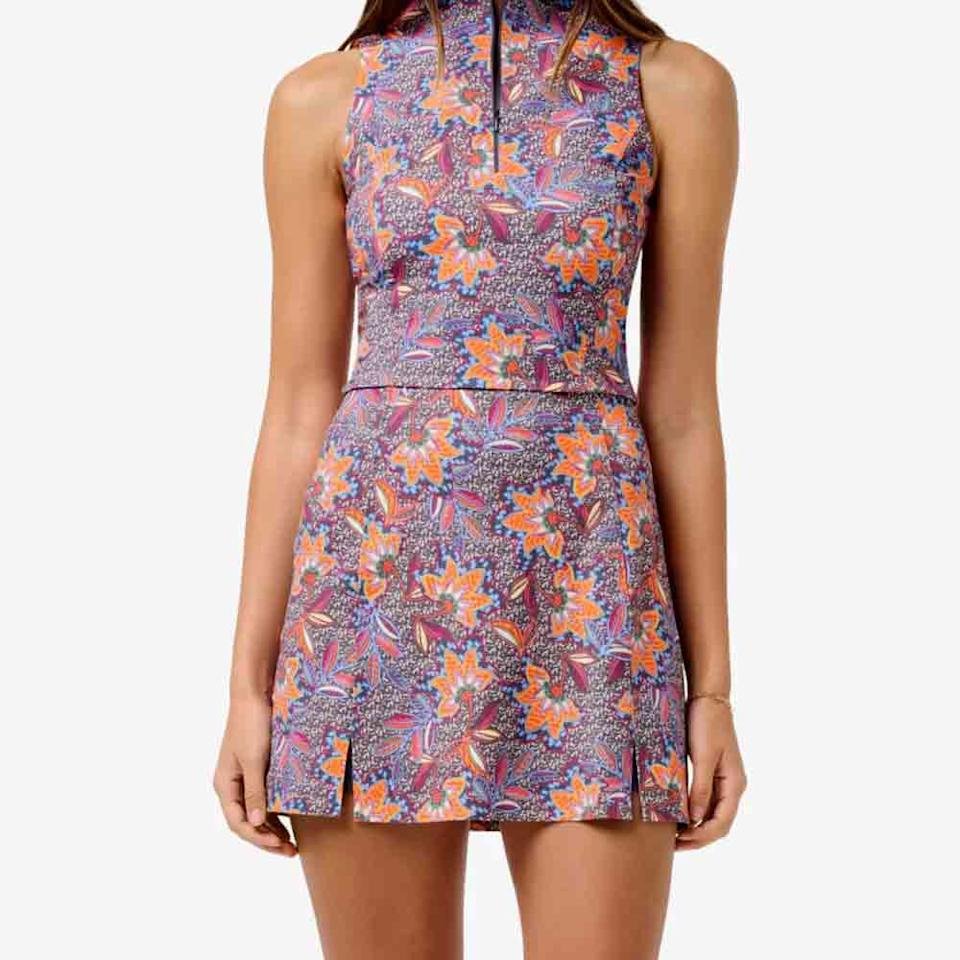 A woman wearing a sleeveless, high-neck, floral dress with a colorful pattern in purple, orange, and pink—a stylish choice for Mother’s Day. The dress features a fitted waist and short hemline with small front slits.