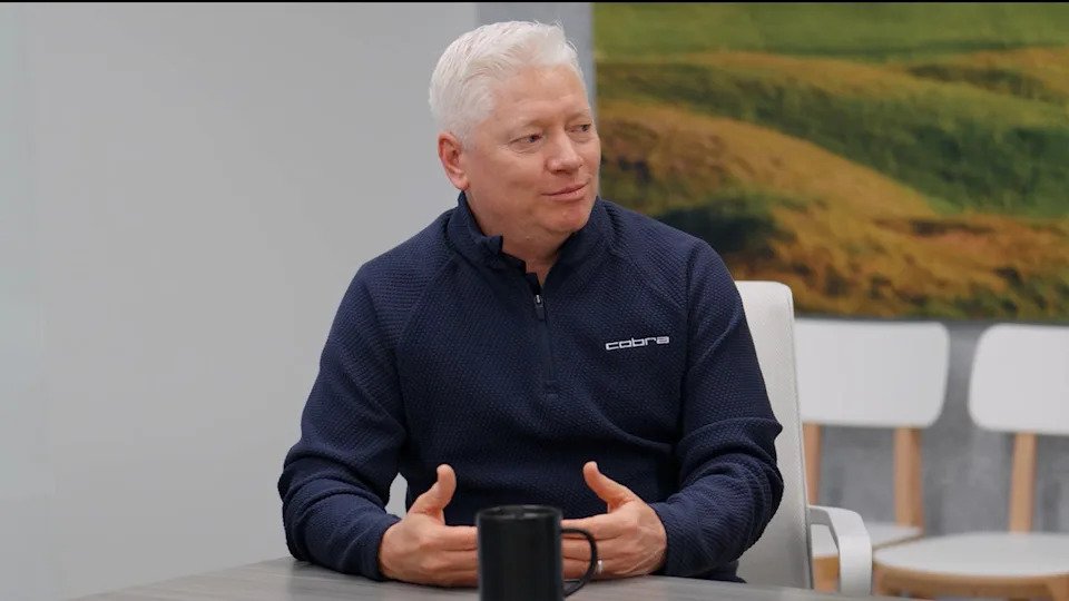 A man with short white hair sits at a table, wearing a dark blue zip-up sweater with Cobra 3DP Irons on it. He gestures as he talks, a black mug in front of him and a blurred landscape visible in the background.