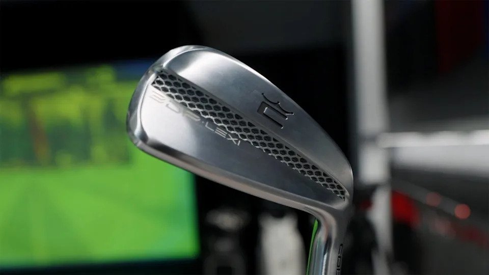 A close-up of a silver Cobra 3DP Iron golf club with a hexagonal pattern and the text 20 DYNAMIC engraved on the club head. The background is blurred with shades of green and black.