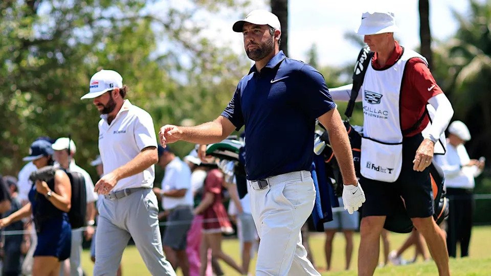 Two golfers walk on a sunny LIV Golf course, each holding clubs, with a caddie in a white bib following behind. Spectators and trees are visible in the background as players react to their shots.